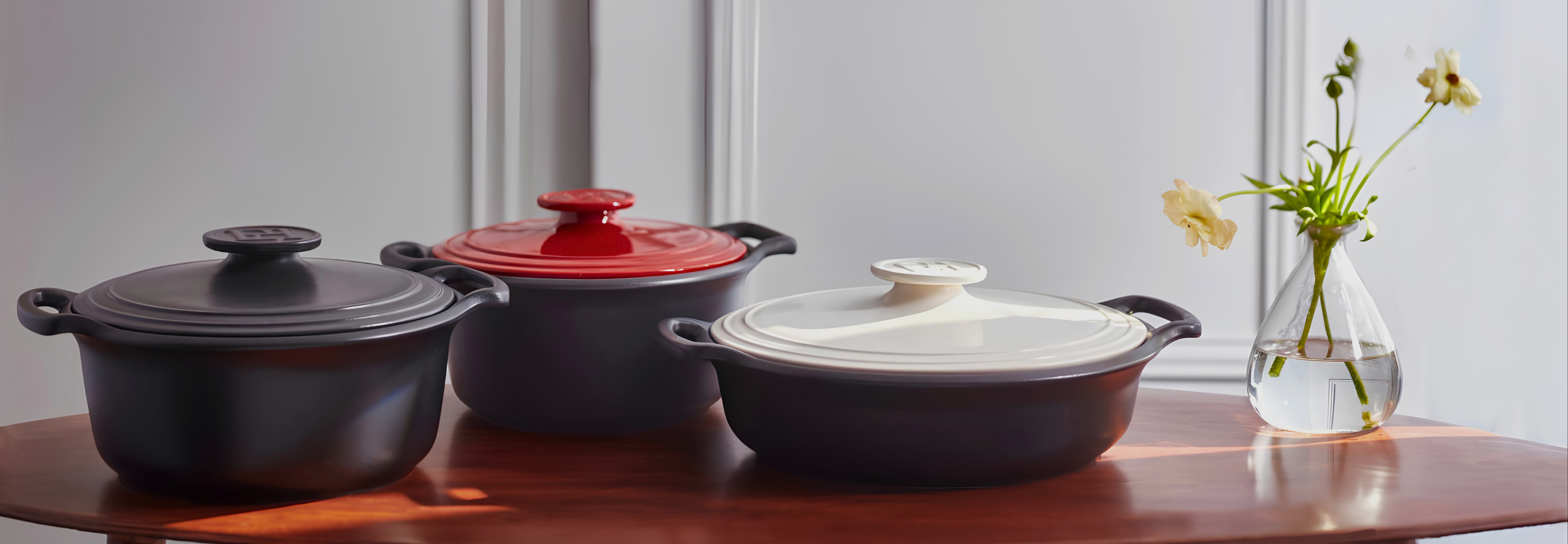 Three ceramic Dutch ovens with lids are arranged on a wooden table in front of a white paneled wall, with a small vase of flowers on the right.